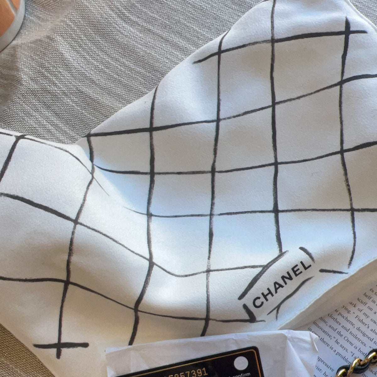 A close-up of a white scarf with a black hand-drawn grid pattern and a visible CHANEL logo patch, laid on a neutral textured surface.