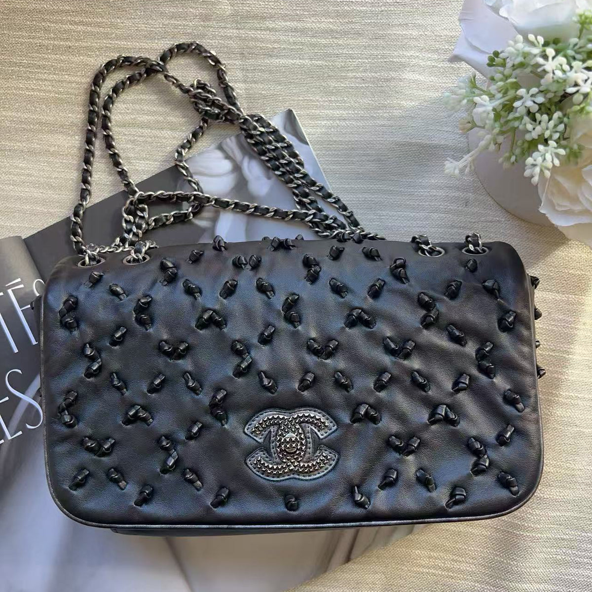 A black quilted Chanel shoulder bag with a rhinestone-embellished interlocking CC logo and a silver-tone chain strap, styled on a beige textured fabric background with a magazine and white flowers.