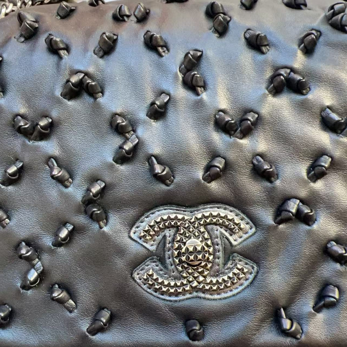 Close-up of a black quilted leather handbag covered in dark studs, featuring a prominent Chanel CC logo patch on the front and a silver-tone chain strap.