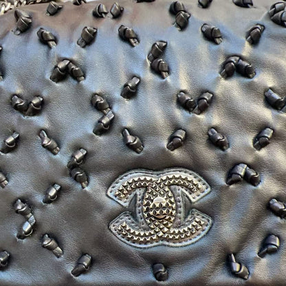 Close-up of a black quilted leather handbag covered in dark studs, featuring a prominent Chanel CC logo patch on the front and a silver-tone chain strap.