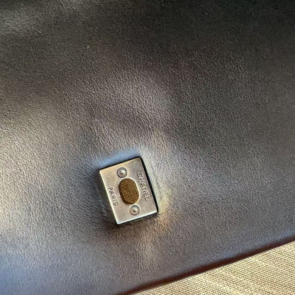 A close-up of a small silver-toned metal square hardware piece with a circular gold center, placed on a dark brown textured leather surface with a beige woven fabric visible in the lower right corner.