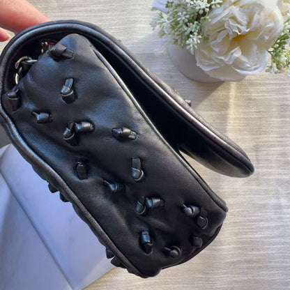 A black leather handbag with a unique textured, quilted top panel covered in small raised studs, shown from a top-down angle on a light wood surface with white flowers and a white box nearby.