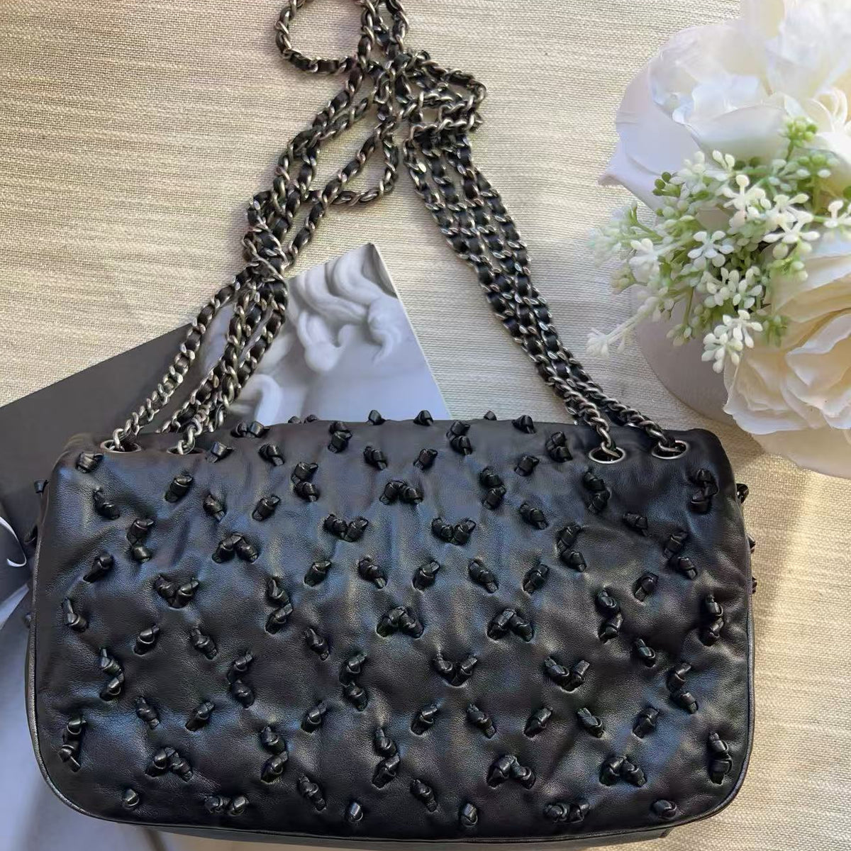 A black quilted leather shoulder bag with a chain strap, decorated with small black bow embellishments, displayed on a light wood surface with white flowers and a magazine in the background.