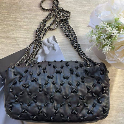 A black quilted leather shoulder bag with a chain strap, decorated with small black bow embellishments, displayed on a light wood surface with white flowers and a magazine in the background.