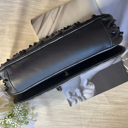 A black leather clutch bag with black metal studs along the top edge, shown from a top-down angle on a light wood-textured surface with a magazine and white flowers as props.