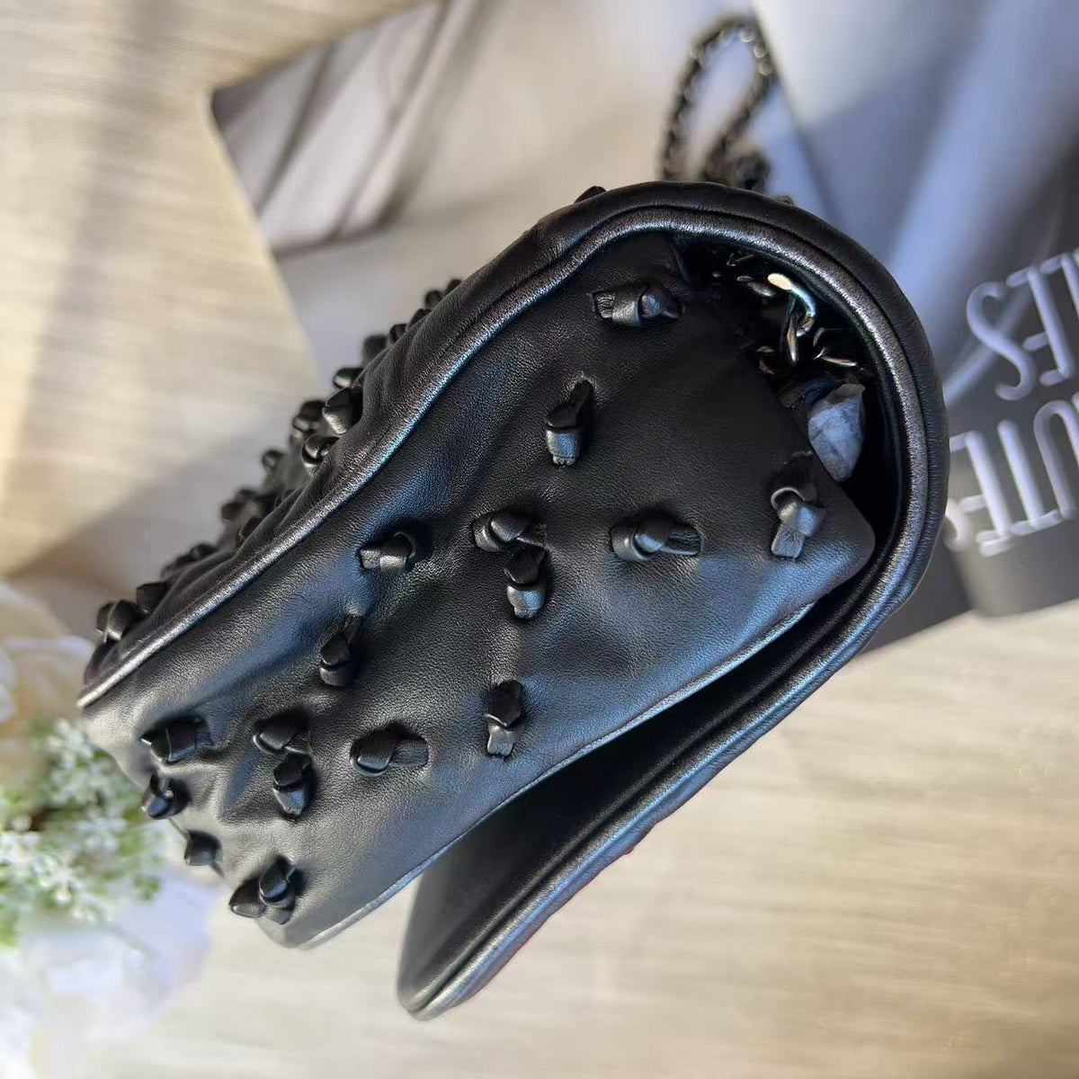 A black leather handbag with prominent black heart-shaped studs, photographed from an overhead angle on a light wood surface next to a gray magazine.