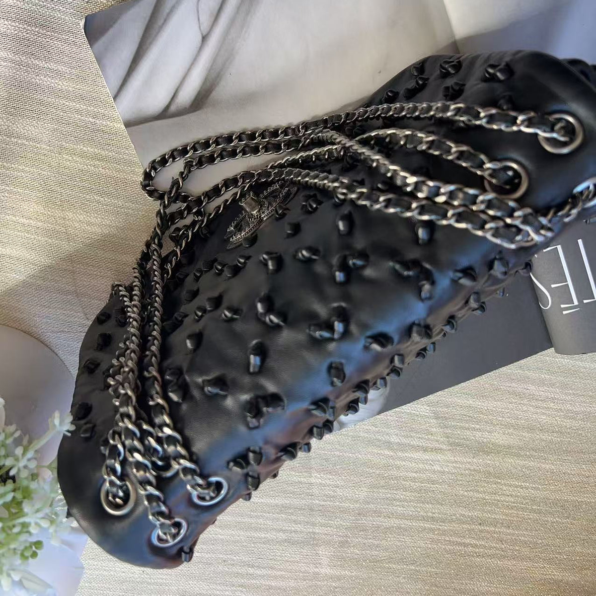 A black textured leather shoulder bag with a quilted pattern, studded details, and silver-tone chain straps, displayed on a light-colored fabric surface.