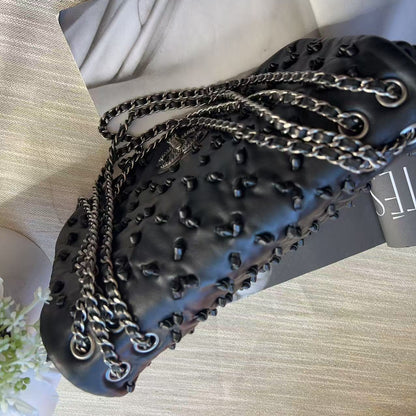 A black textured leather shoulder bag with a quilted pattern, studded details, and silver-tone chain straps, displayed on a light-colored fabric surface.