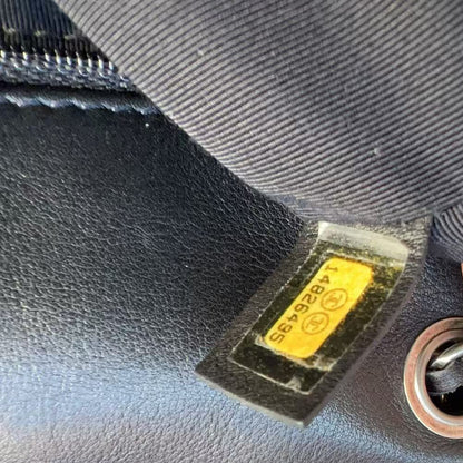 Close-up of a black leather handbag showing a yellow Chanel logo patch, metal chain detail, and a finger holding the fabric.