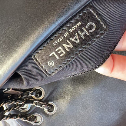 Close-up of a black Chanel handbag showing the interior lining and a stitched black label with white CHANEL text and 'MADE IN ITALY'. A finger is holding the bag open, and part of a black-and-silver chain strap is visible.