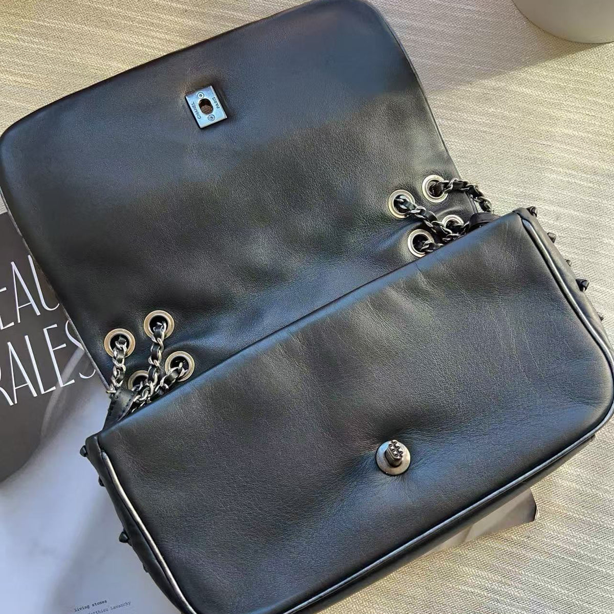 A black leather shoulder bag with silver-tone chain details and hardware, photographed from above on a light wood-textured surface next to a magazine.