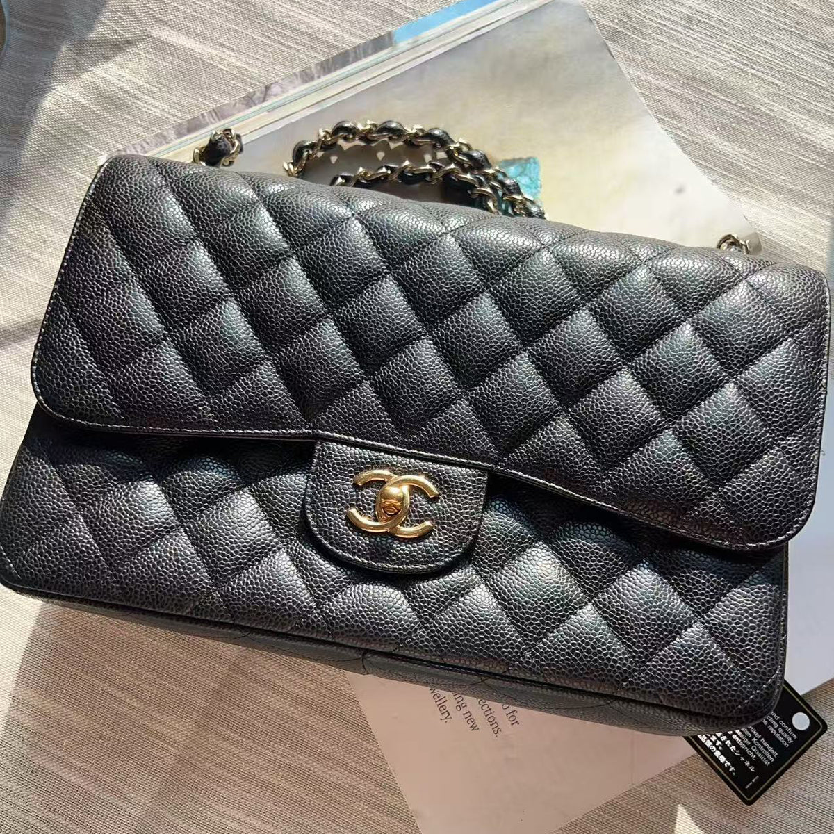 A black quilted leather handbag with a gold-tone interlocking CC clasp, resting on a light-colored surface with its original paper tag and packaging visible.
