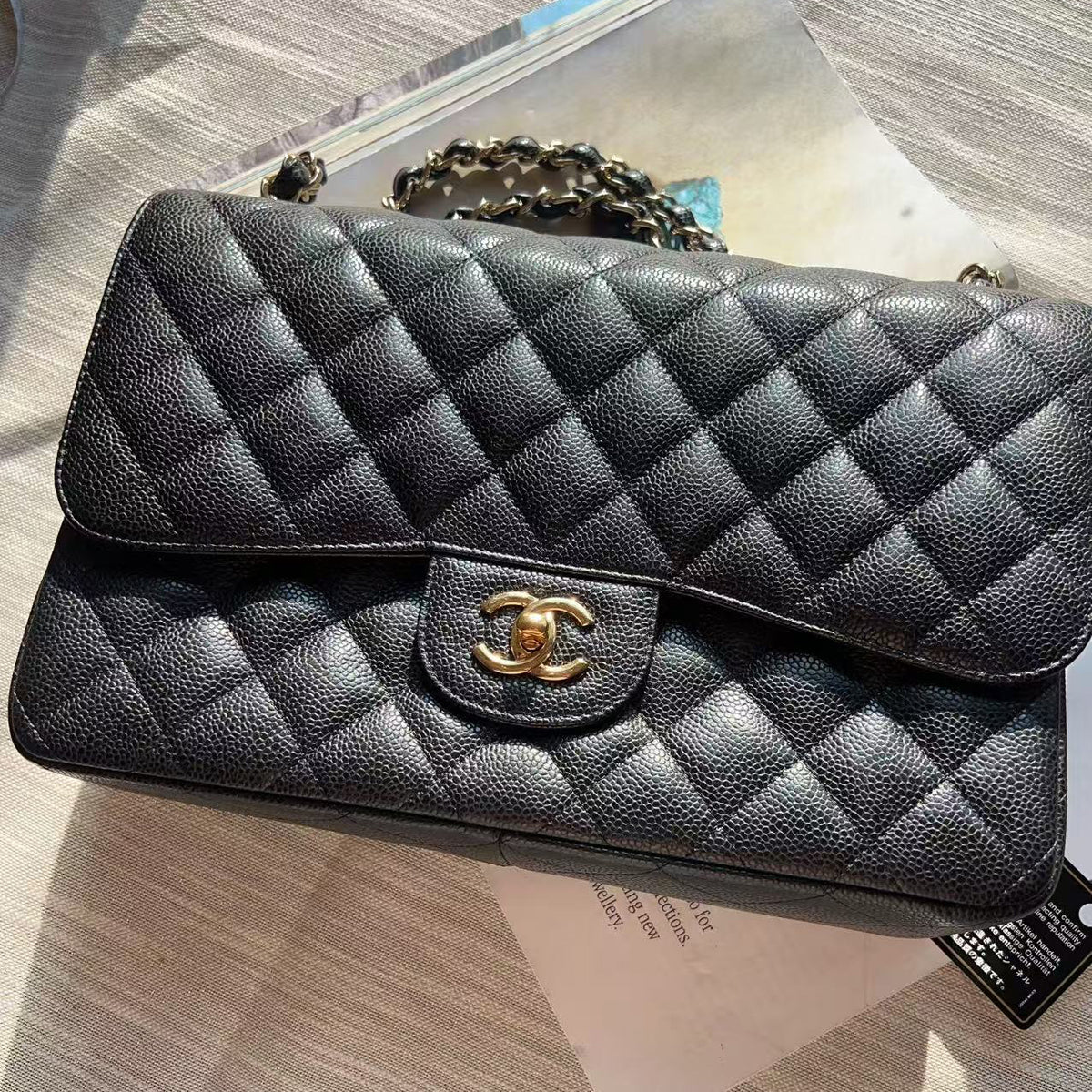 A black quilted leather handbag with a gold-tone interlocking CC logo clasp, resting on a light-colored surface with papers underneath.