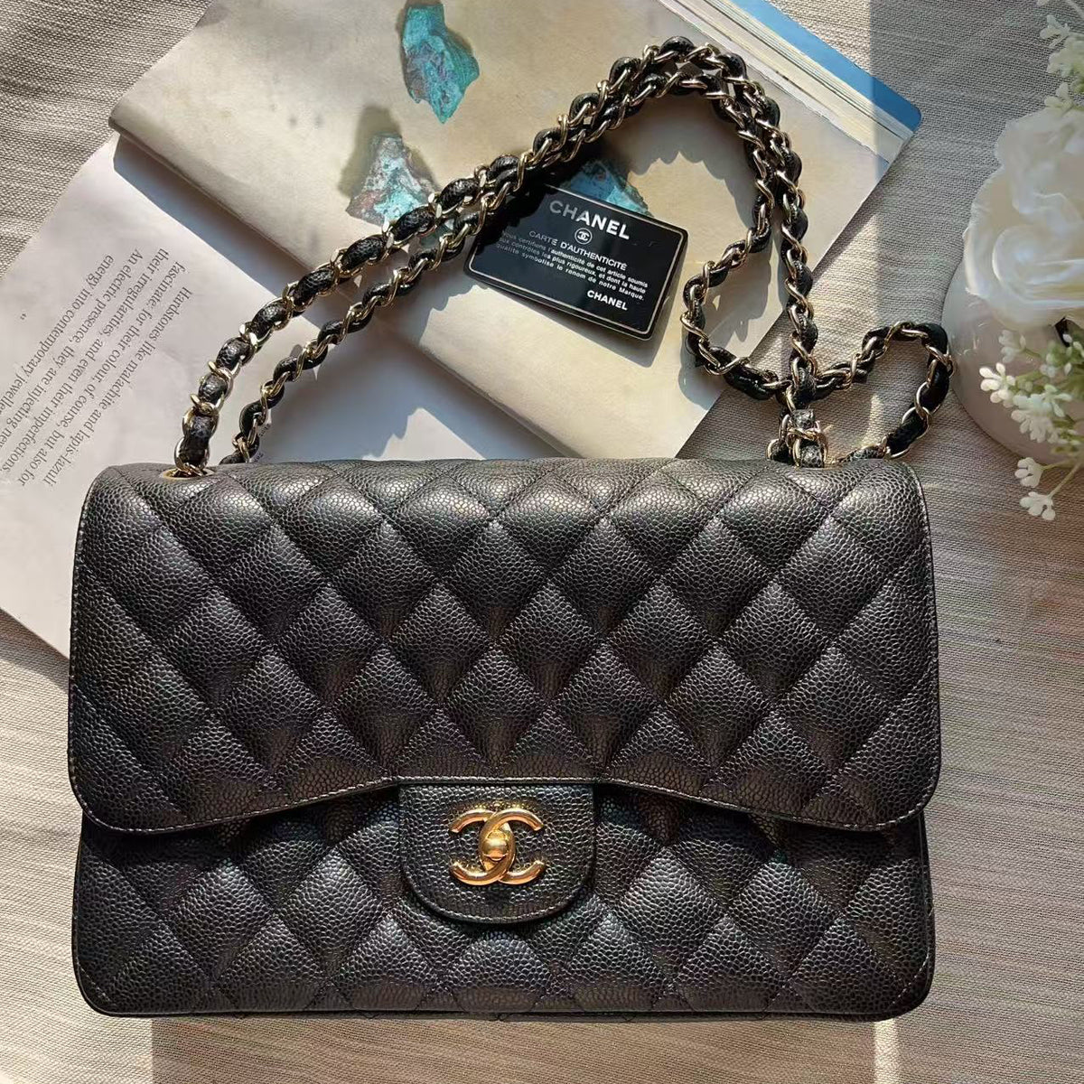 A black quilted Chanel flap shoulder bag with a gold-tone CC turn-lock closure and chain strap, laid flat on a light wood surface with an open magazine and small flowers in the background.