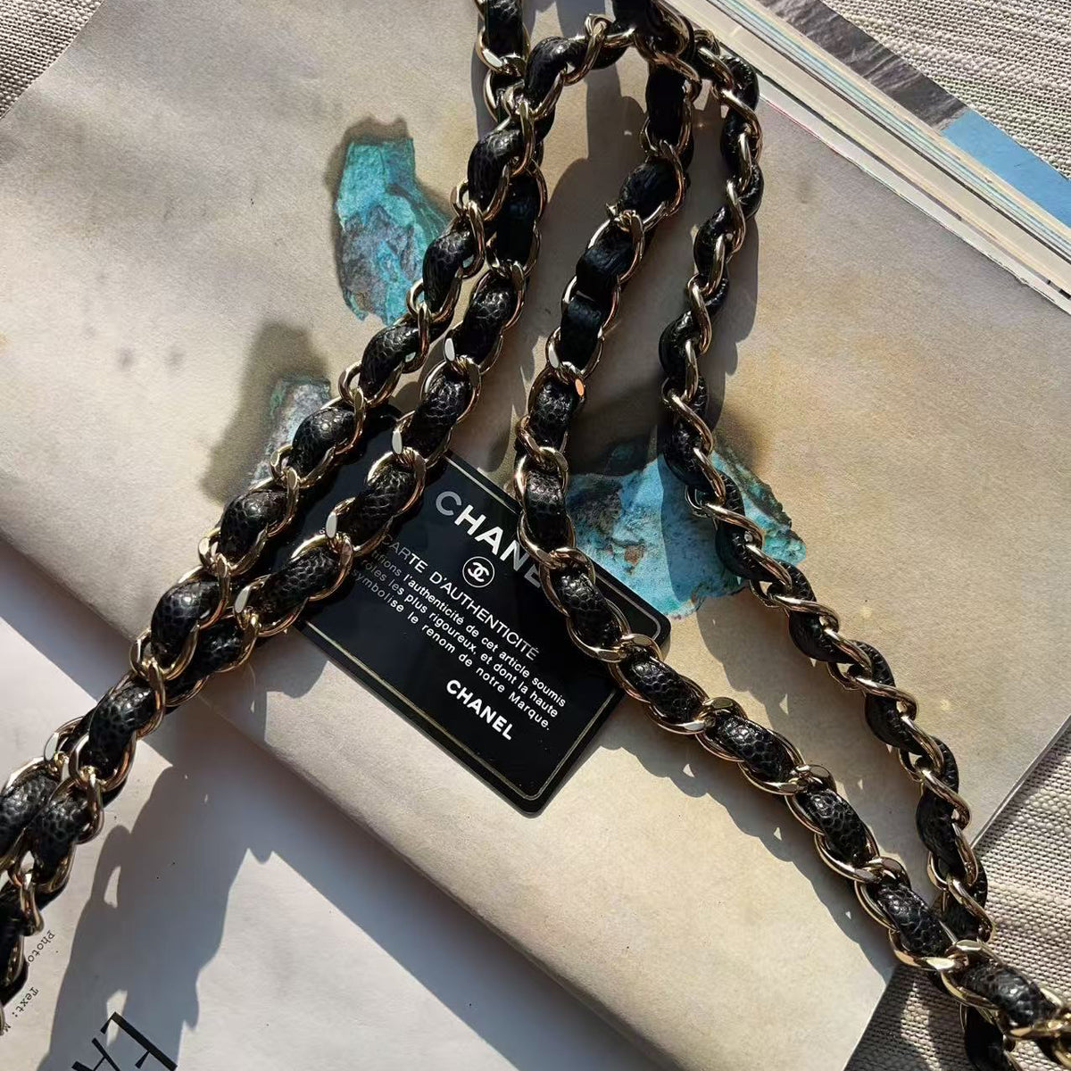 A close-up of a Chanel shoulder bag strap made of black leather and gold-tone chain, laid over a magazine page with a visible Chanel hangtag.