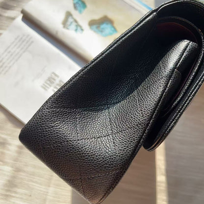 A close-up of a black pebbled leather clutch or wallet, angled diagonally on a light wood surface with an open book in the background.