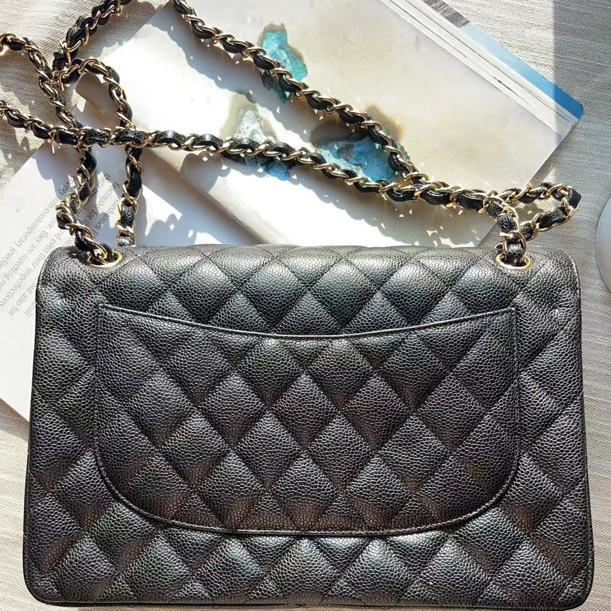 A black quilted leather shoulder bag with a gold-tone chain strap, laid flat on a light-colored wooden surface next to a magazine.