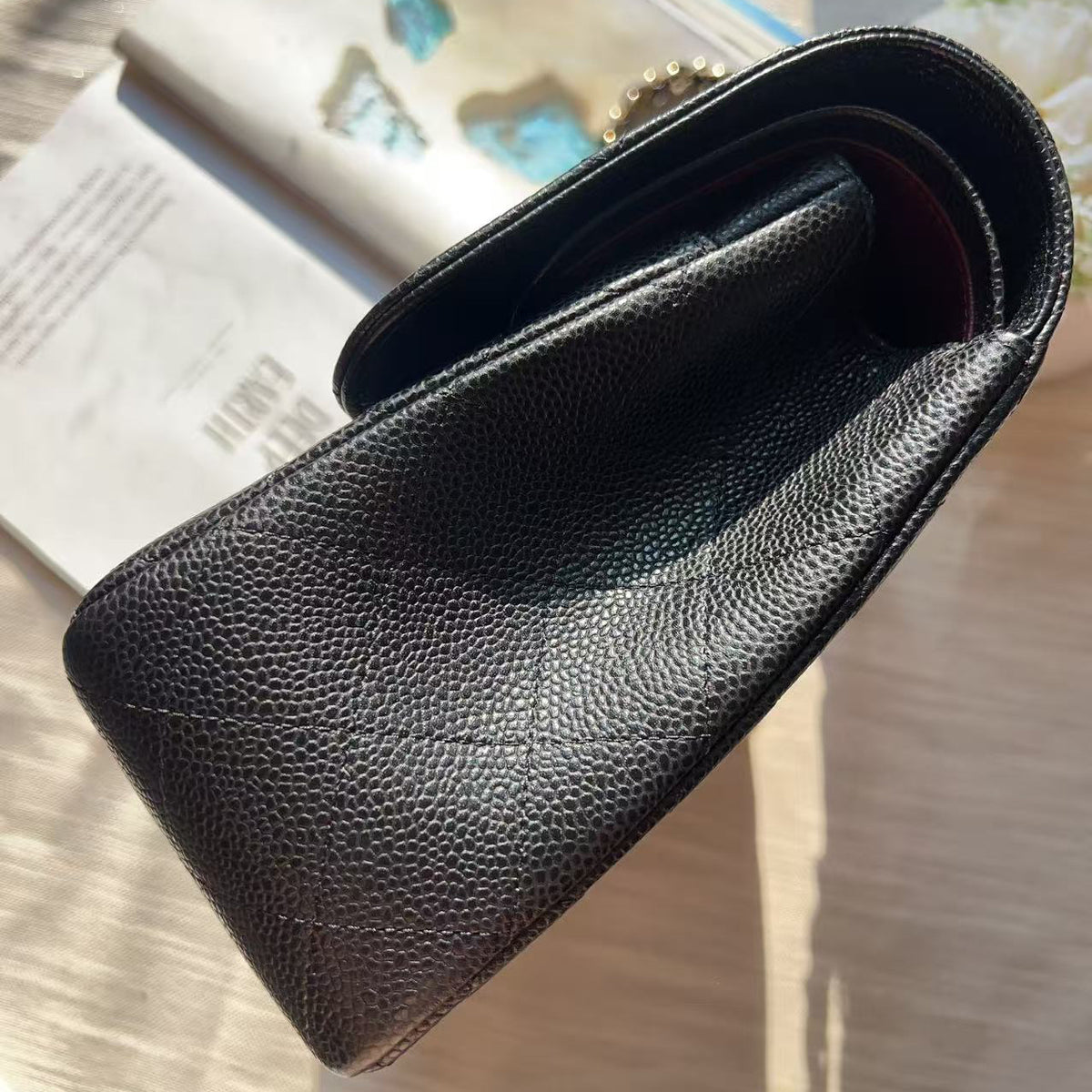 A black pebbled leather clutch bag with a gold-tone ring pull, shown from an overhead angle on a light wood surface next to an open magazine.