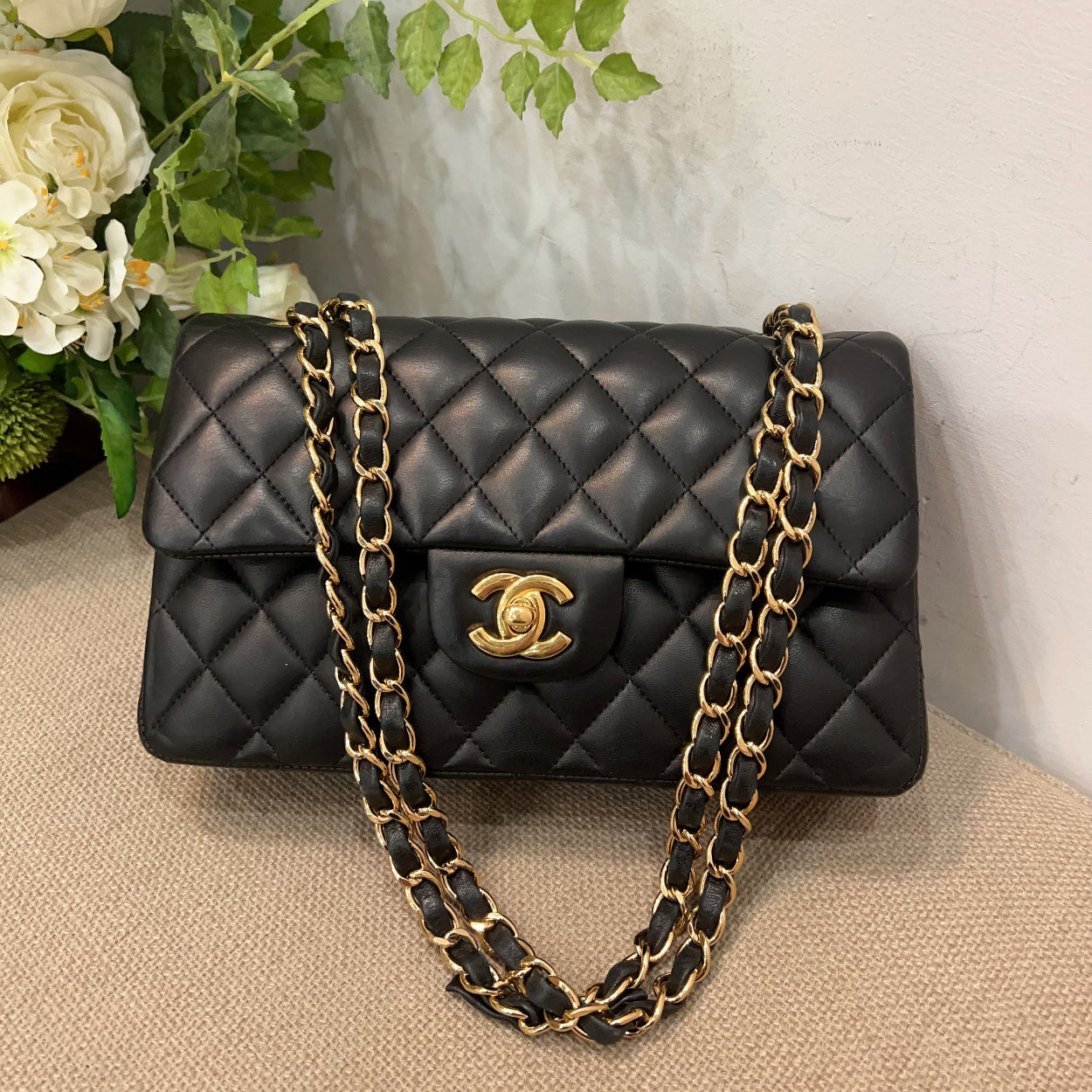 Black quilted handbag with gold chain on a beige surface with flowers in the background