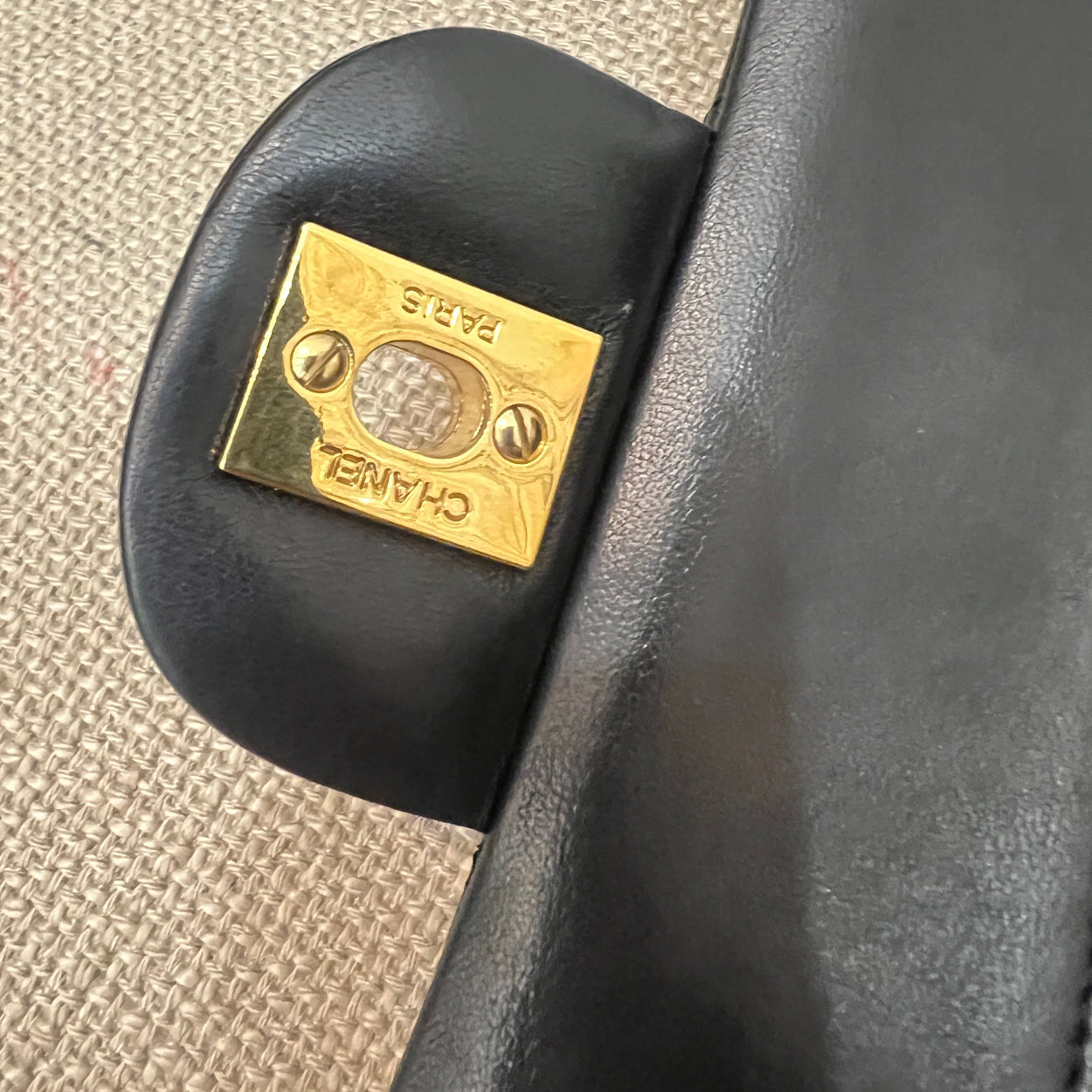 Close-up of a black handbag with a gold Chanel logo on a textured beige surface.