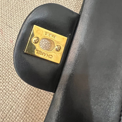 Close-up of a black handbag with a gold Chanel logo on a textured beige surface.