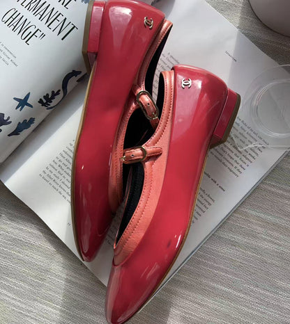 A pair of glossy red Mary Jane flats with pointed toes and low block heels, styled on top of an open magazine over a gray textured surface.