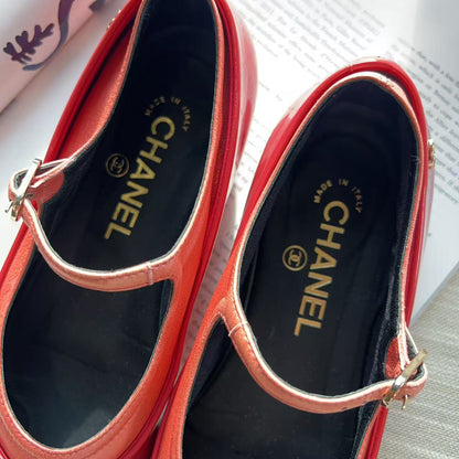 A pair of red Chanel Mary Jane shoes with gold-tone buckle straps, displayed on a light-colored surface with printed papers in the background.