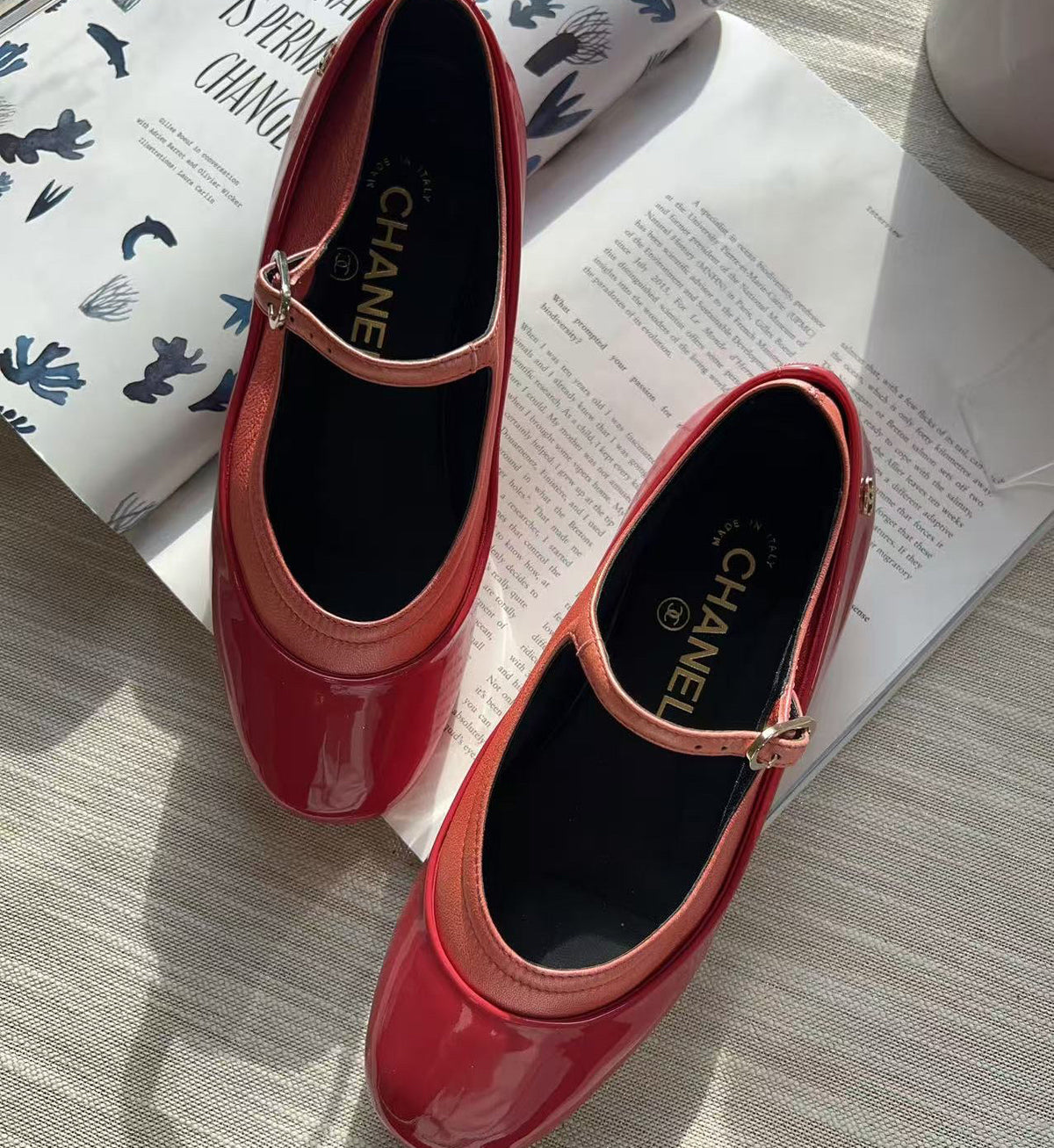 A pair of glossy red patent leather Mary Jane shoes with a small gold buckle, displayed on a light wood surface next to an open magazine. The brand name CHANEL is visible on the insoles.