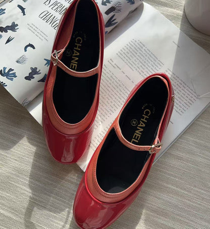 A pair of glossy red patent leather Mary Jane shoes with a small gold buckle, displayed on a light wood surface next to an open magazine. The brand name CHANEL is visible on the insoles.
