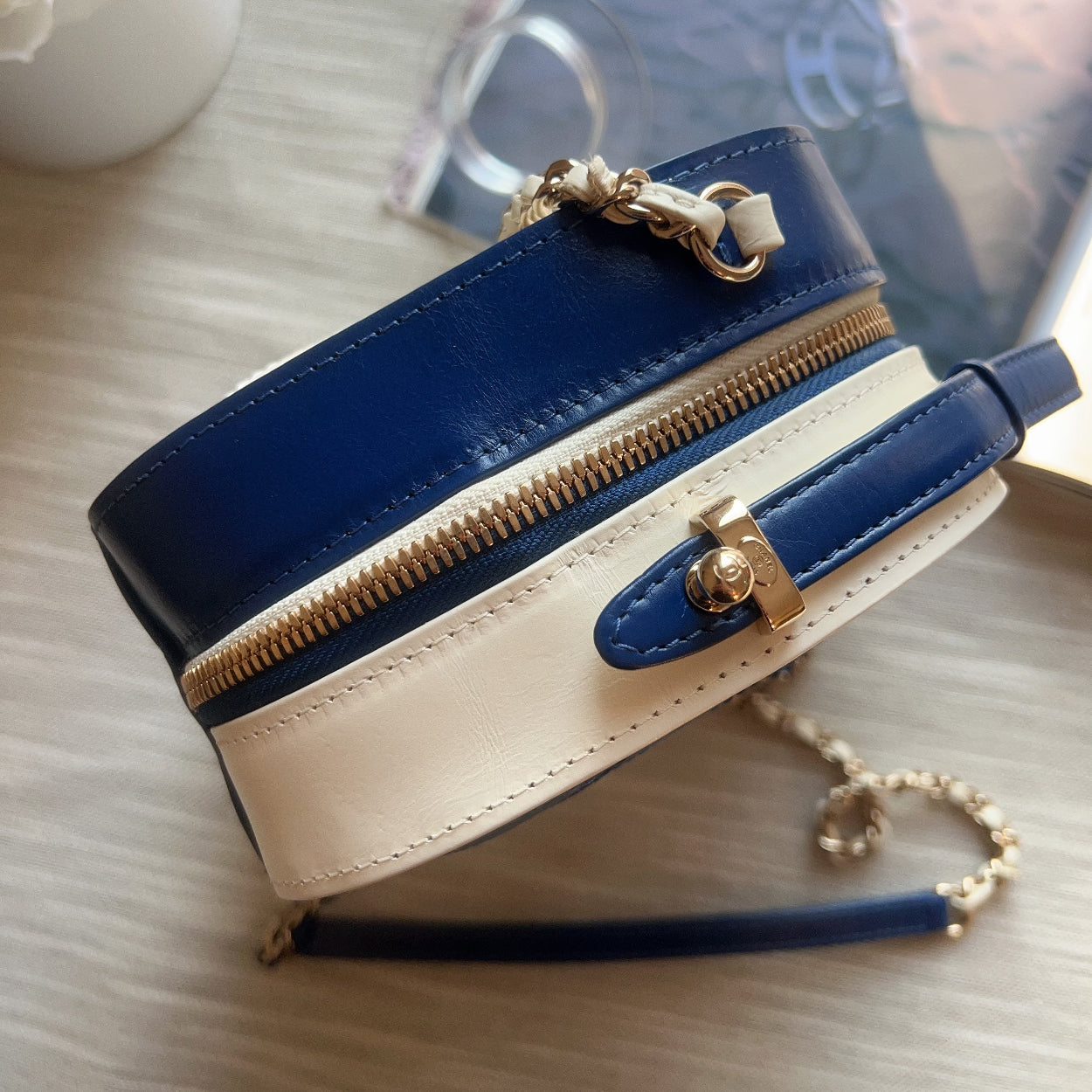 Blue and beige handbag with gold zipper and hardware on a light wooden surface.