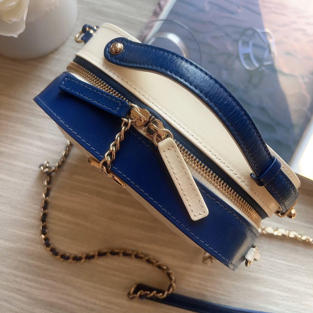 Blue and beige handbag with gold chain on a light wooden surface.
