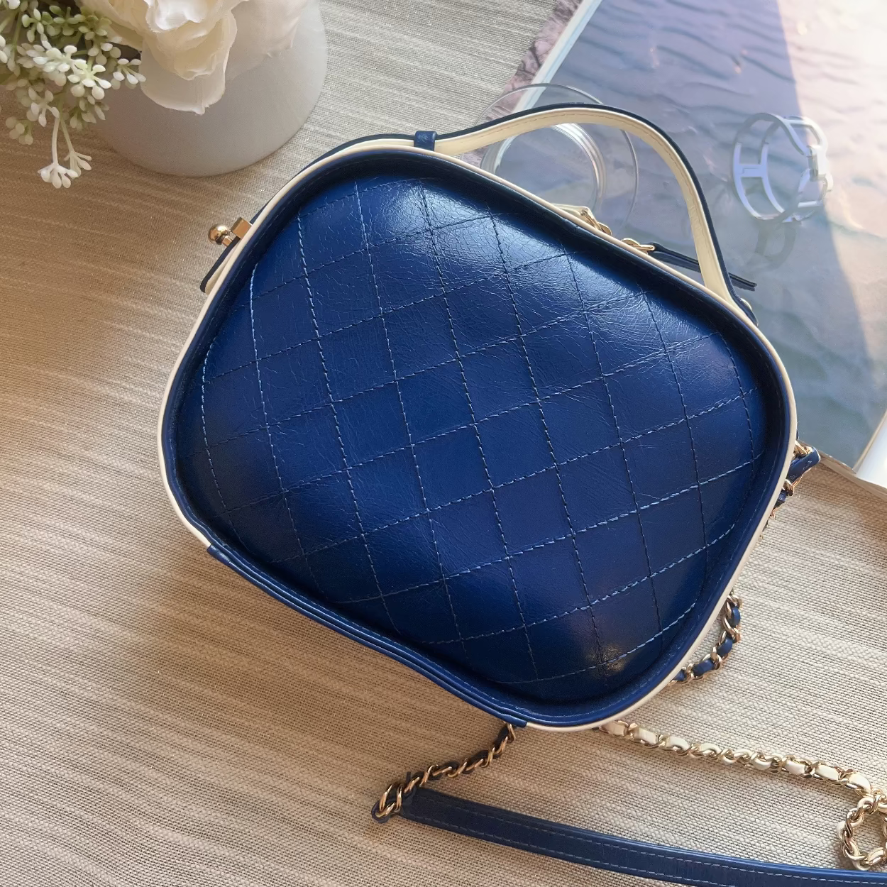 Blue quilted handbag on a wooden surface with a vase of flowers and a tablet in the background.