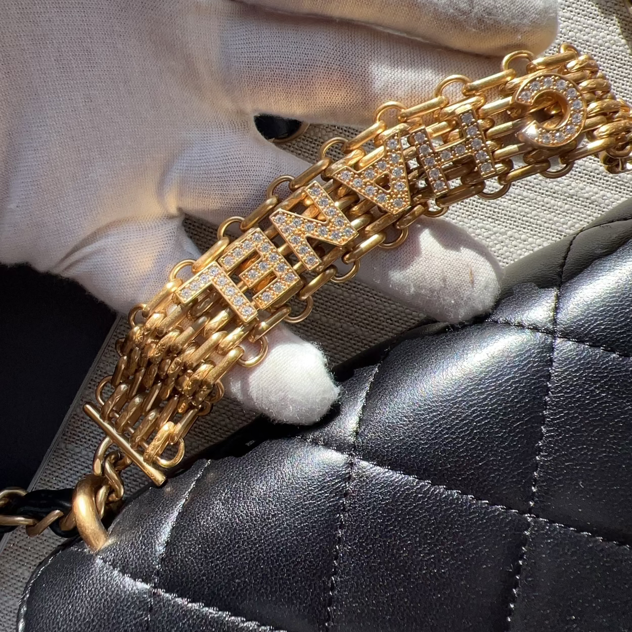 Gold bracelet with 'FENDI' branding held by a hand against a black quilted surface.