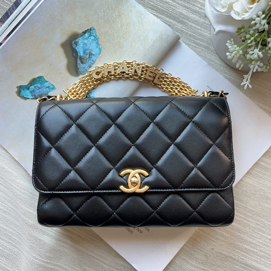 A black quilted leather shoulder bag with a gold-tone interlocking CC logo and a gold chain strap, styled on top of a magazine over a light beige fabric background.