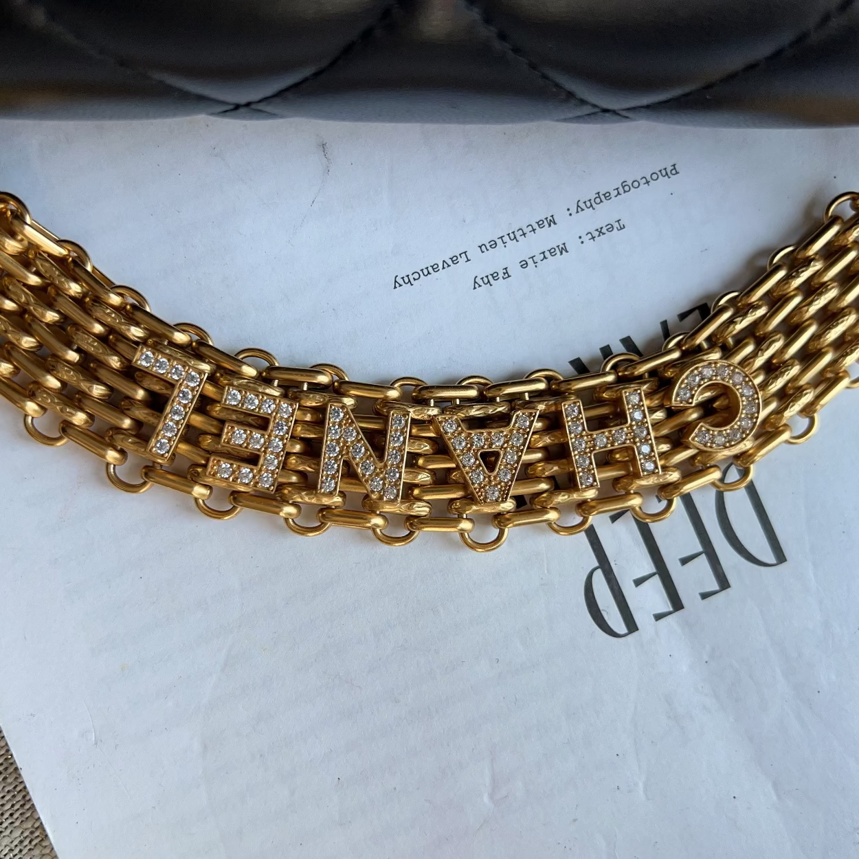 A gold-tone chain bracelet with the word 'CHANEL' spelled out in rhinestone-encrusted letters, displayed on a white printed sheet.
