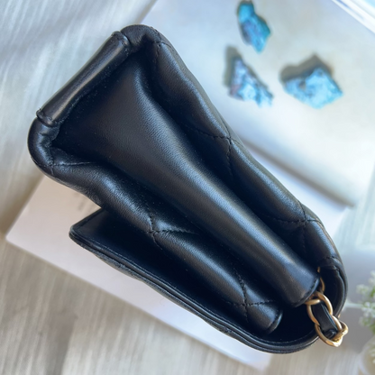 A black quilted leather pouch with a gold-tone metal ring clasp, styled on a white book over a light wood surface with a floral prop.