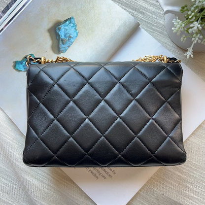 A black quilted leather shoulder bag with a gold chain strap, styled on top of an open book with decorative blue stones and white flowers.