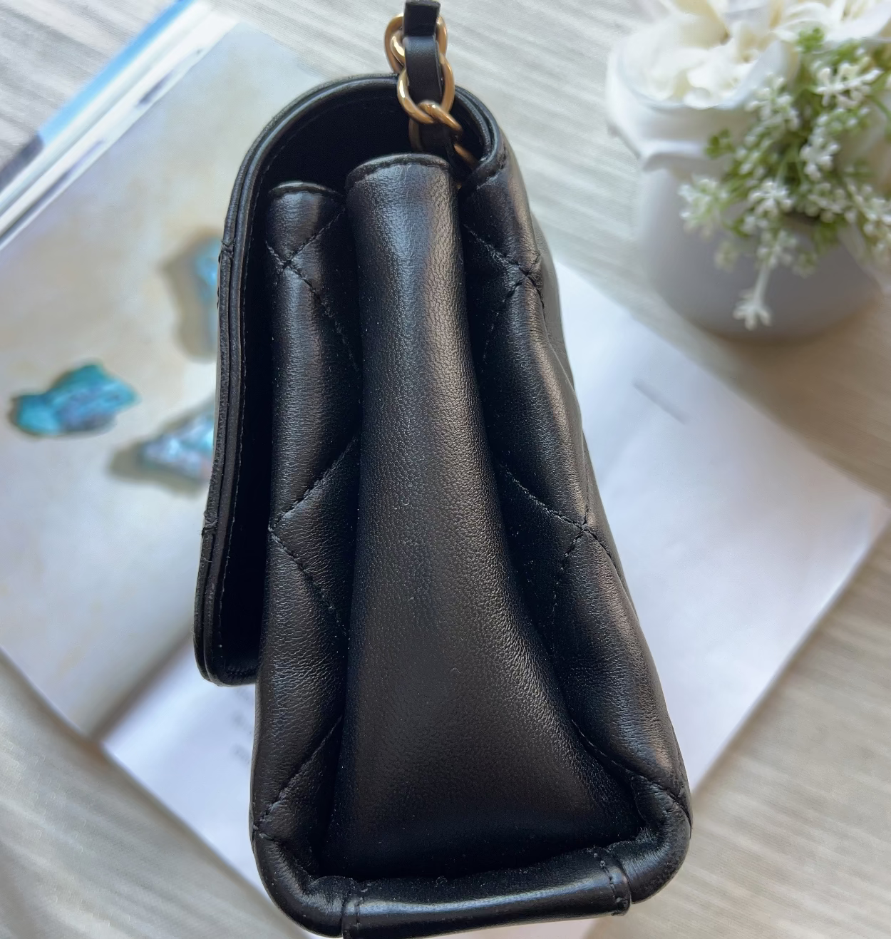 A black quilted leather handbag shown from a top-down angle, resting on a white book or magazine over a light wood surface, with a small white flower in the corner.