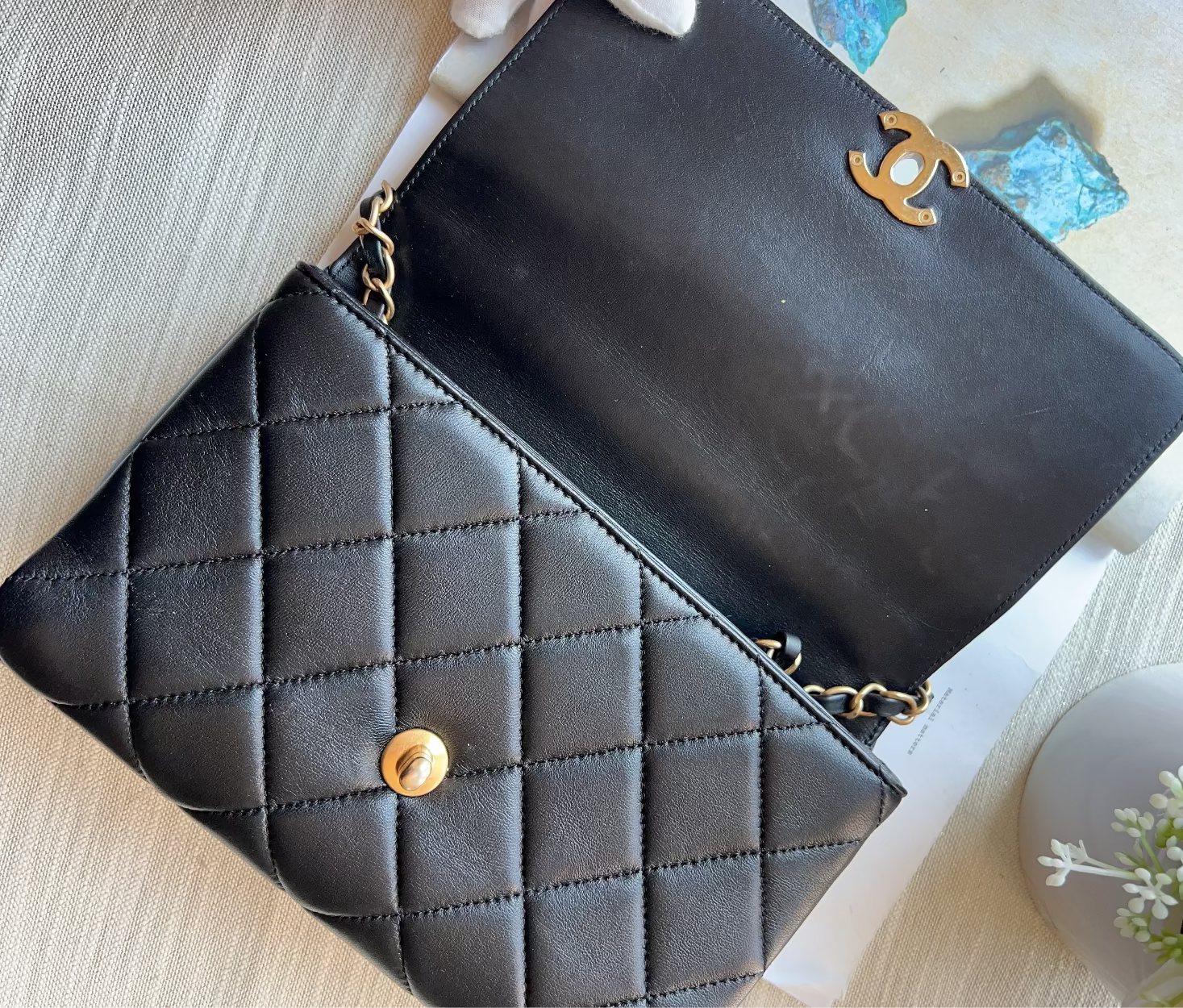 A black quilted leather shoulder bag with gold-tone hardware, shown open on a light beige textured surface. The bag features a diamond-quilted pattern on the front flap with a gold snap button, and a gold interlocking CC logo on the inside flap, identifying it as Chanel.