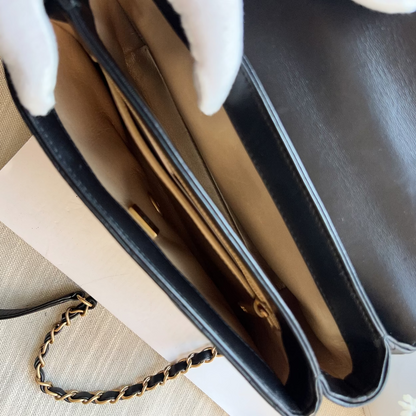 Close-up of a black leather handbag interior with a gold-tone chain strap, shown with white-gloved hands holding the bag open on a light beige surface.