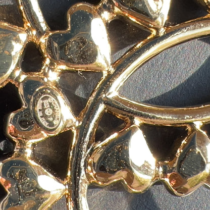 Close-up of a gold-toned metal brooch with a floral or butterfly-like openwork design, showing reflective polished surfaces against a dark background.