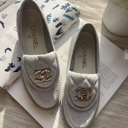 A pair of Chanel loafers in light gray suede with gold-tone interlocking CC logos, displayed on an open book with a folded patterned scarf in the background.
