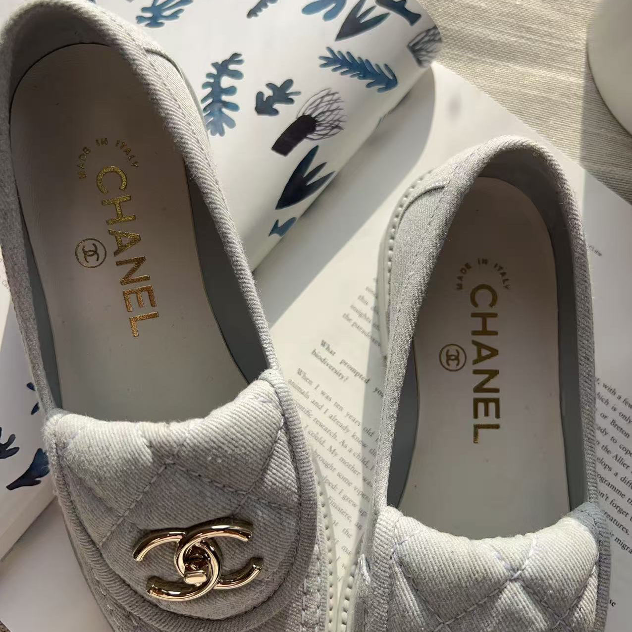 A pair of Chanel slip-on loafers in light gray quilted canvas with gold-tone interlocking CC logos, displayed on top of printed papers and a patterned bag.