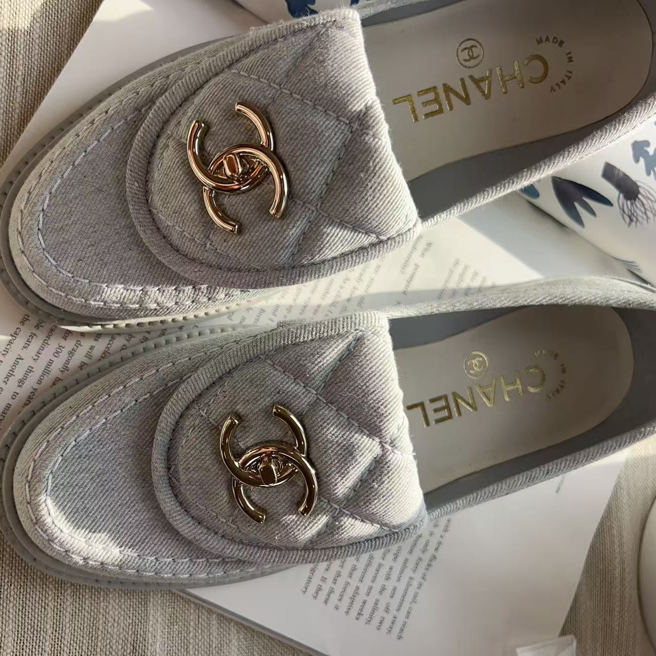 A pair of Chanel loafers in light gray quilted canvas with gold-tone interlocking CC logos, displayed on a white book over a beige textured fabric surface.