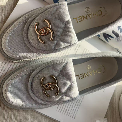 A pair of Chanel loafers in light gray quilted canvas with gold-tone interlocking CC logos, displayed on a white book over a beige textured fabric surface.