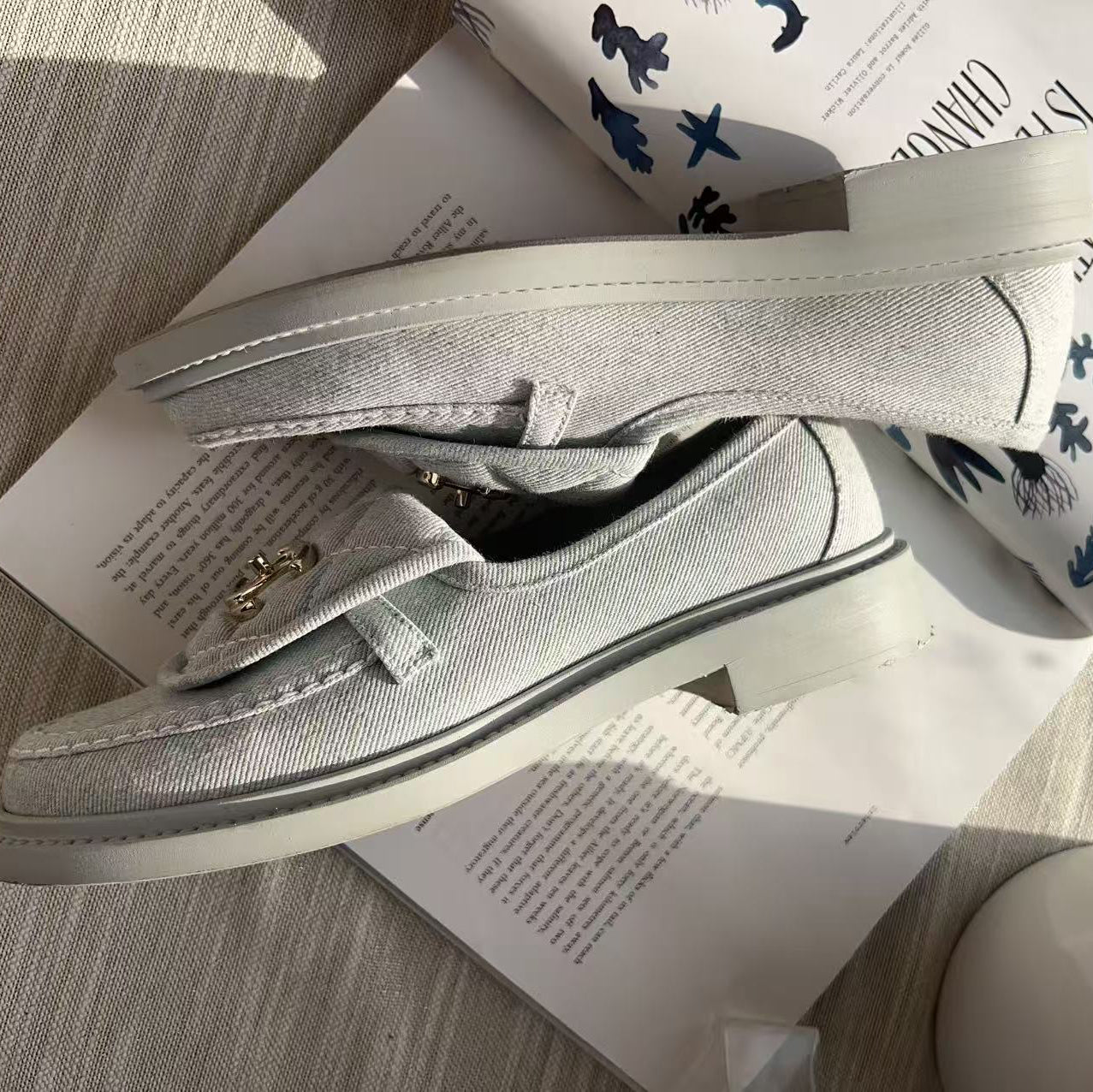A pair of light gray canvas slip-on loafers with a small gold-tone metal accent on the strap, displayed on top of printed books on a textured gray surface.