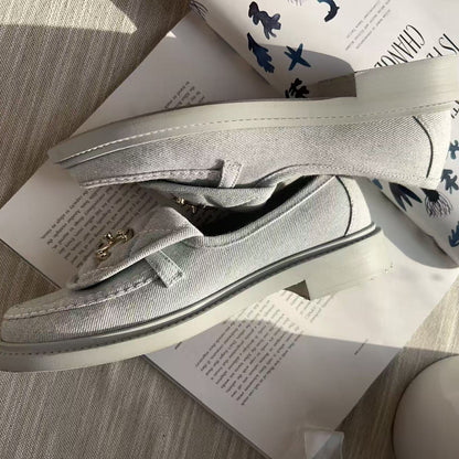 A pair of light gray canvas slip-on loafers with a small gold-tone metal accent on the strap, displayed on top of printed books on a textured gray surface.