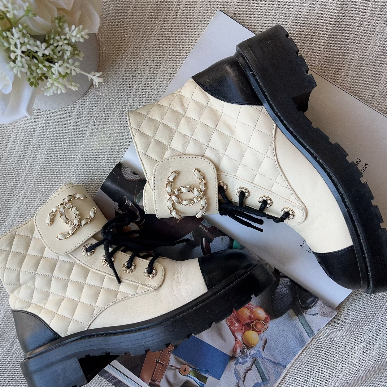 Pair of quilted leather boots with a brand logo on a textured surface with flowers and a magazine.