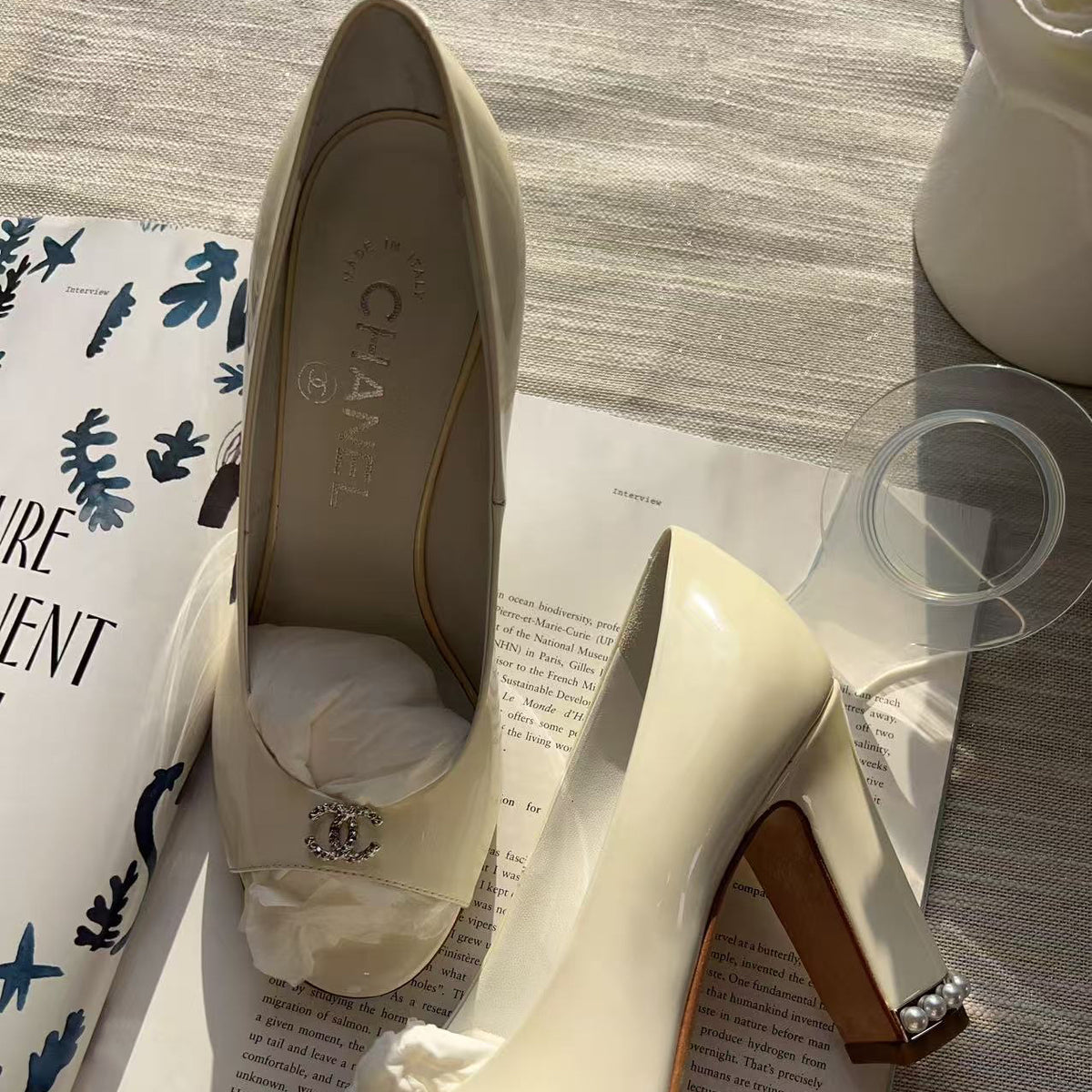 A pair of Chanel high-heeled pumps in off-white patent leather, styled on top of an open book with a Chanel box visible on the left. The shoes feature a block heel, a small crystal-embellished detail on the toe, and a crystal-accented block heel.