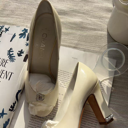 A pair of Chanel high-heeled pumps in off-white patent leather, styled on top of an open book with a Chanel box visible on the left. The shoes feature a block heel, a small crystal-embellished detail on the toe, and a crystal-accented block heel.